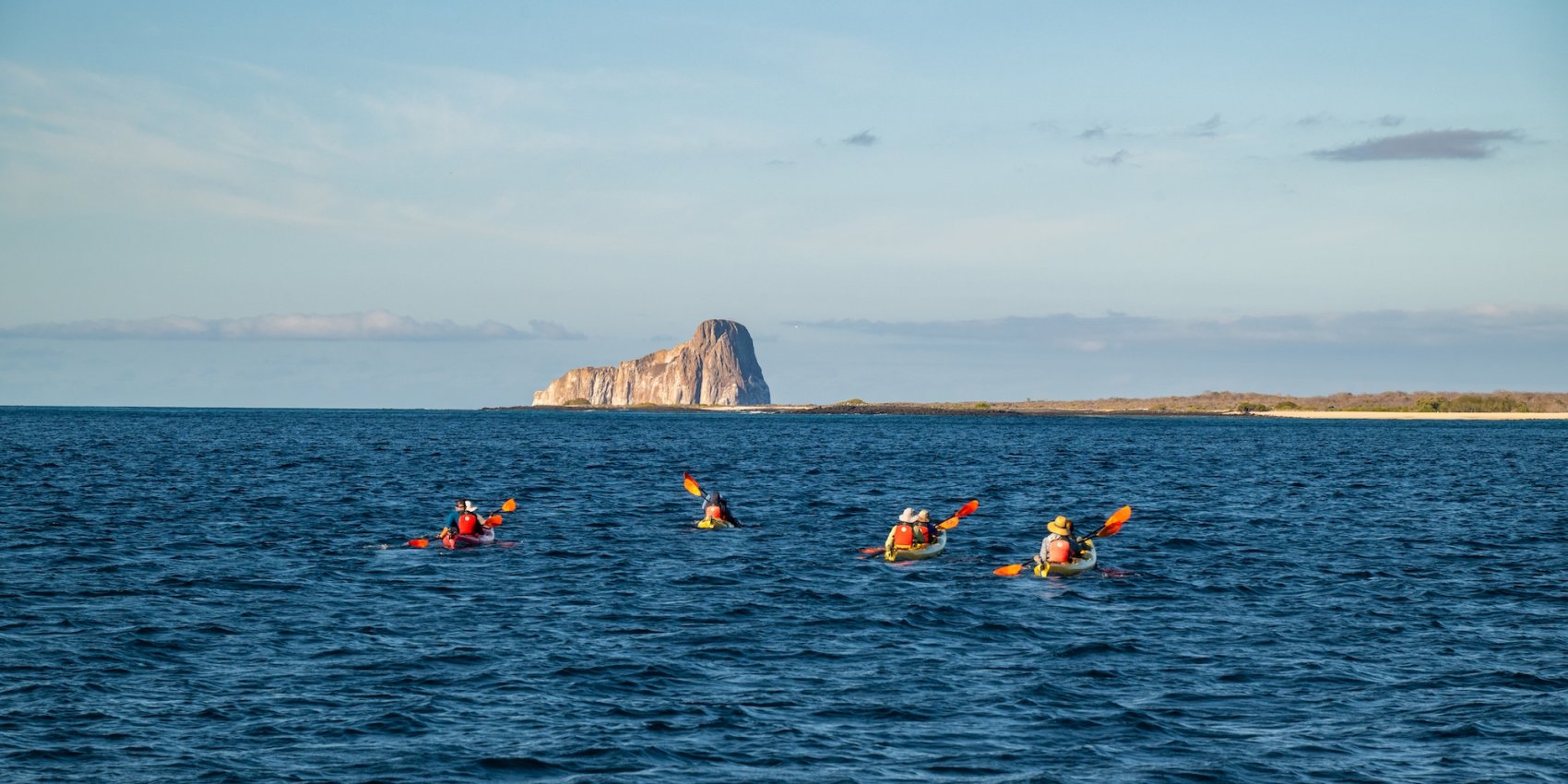 Several kayakers paddle across the ocean toward the rocky islet of León Dormido (Kicker Rock) off the coast of the Galápagos Islands, under a clear blue sky.