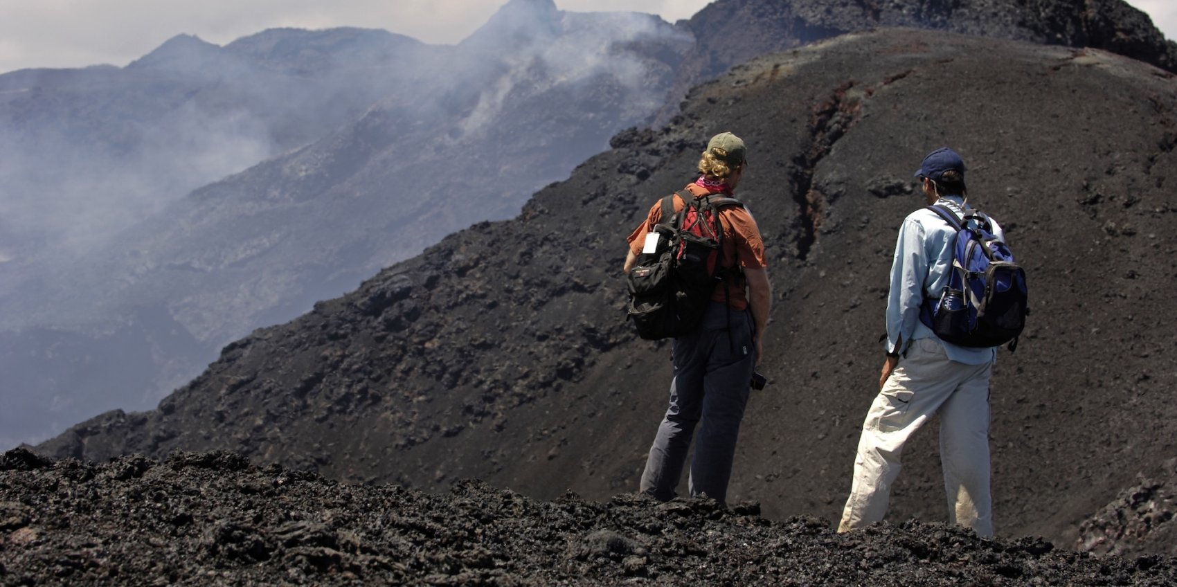 Two hikers stand atop black volcanic terrain in the Galápagos Islands, looking out over a dramatic landscape of active volcanic craters and steaming slopes.